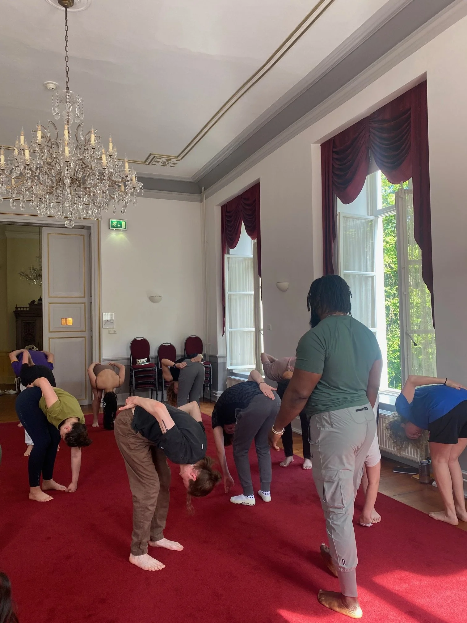 People participating in a yoga or stretching class in a bright room with large windows and a chandelier, some lying on the floor, others standing or bending over.