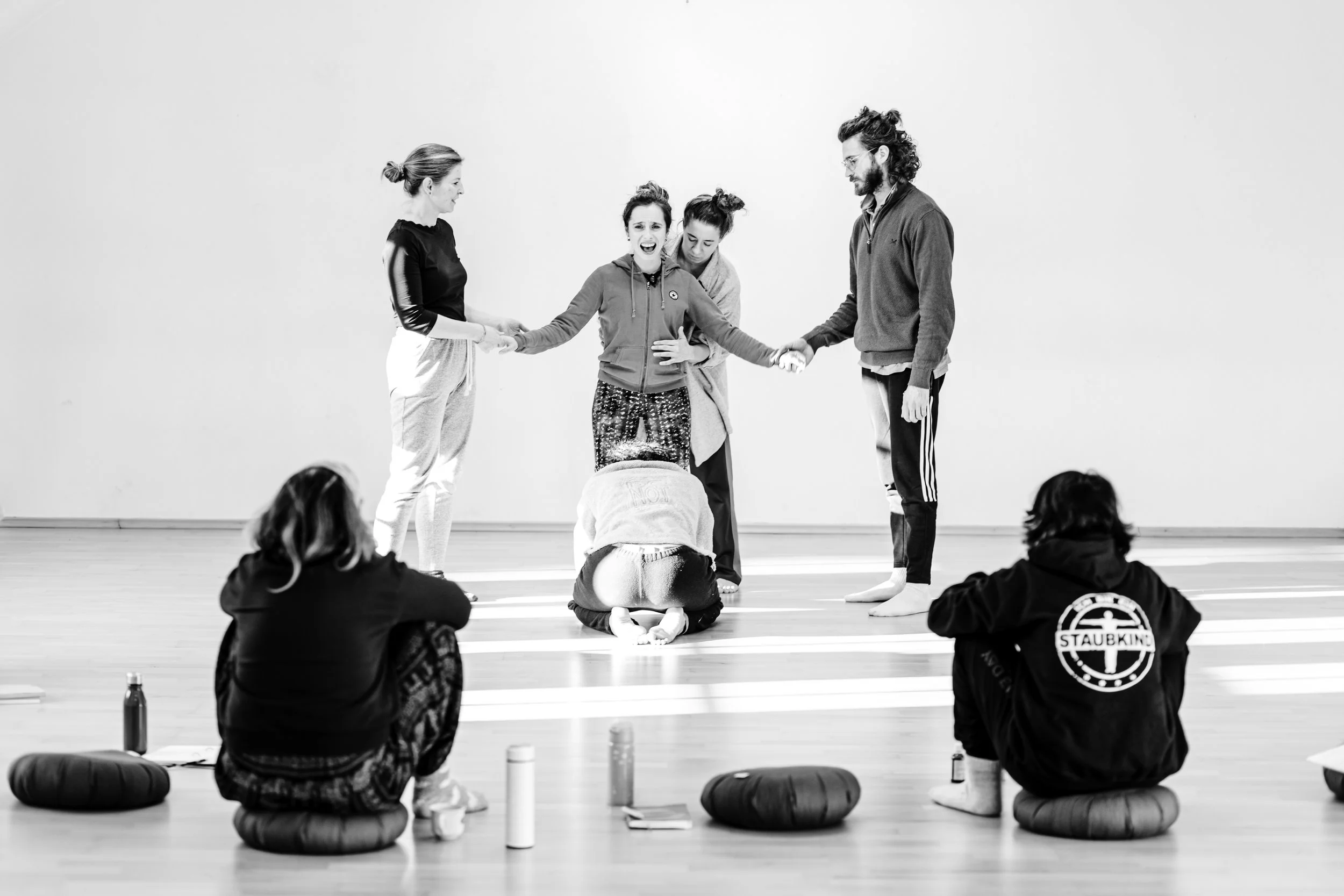 Black and white photo of a dance class or workshop with four people on stage: two women on either side, holding hands, and a woman in the middle with a man on the right. The woman in the middle appears to be emotional, with eyes closed and mouth open. The people in the audience, some seated on floor cushions, are watching the scene.