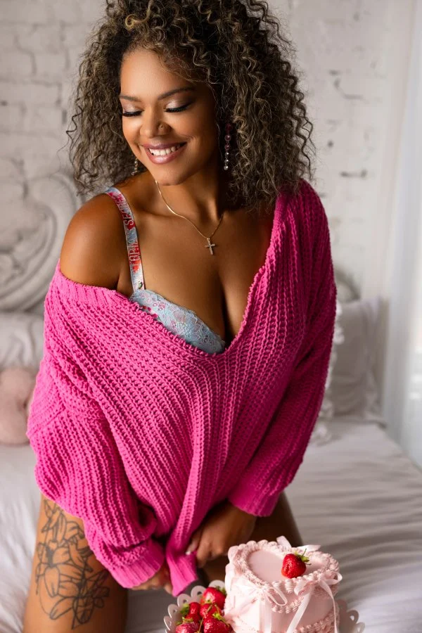 A smiling woman with curly hair wearing a pink sweater and floral bra, sitting on a bed with a pink cake decorated with strawberries in front of her, in a cozy room with white brick walls.