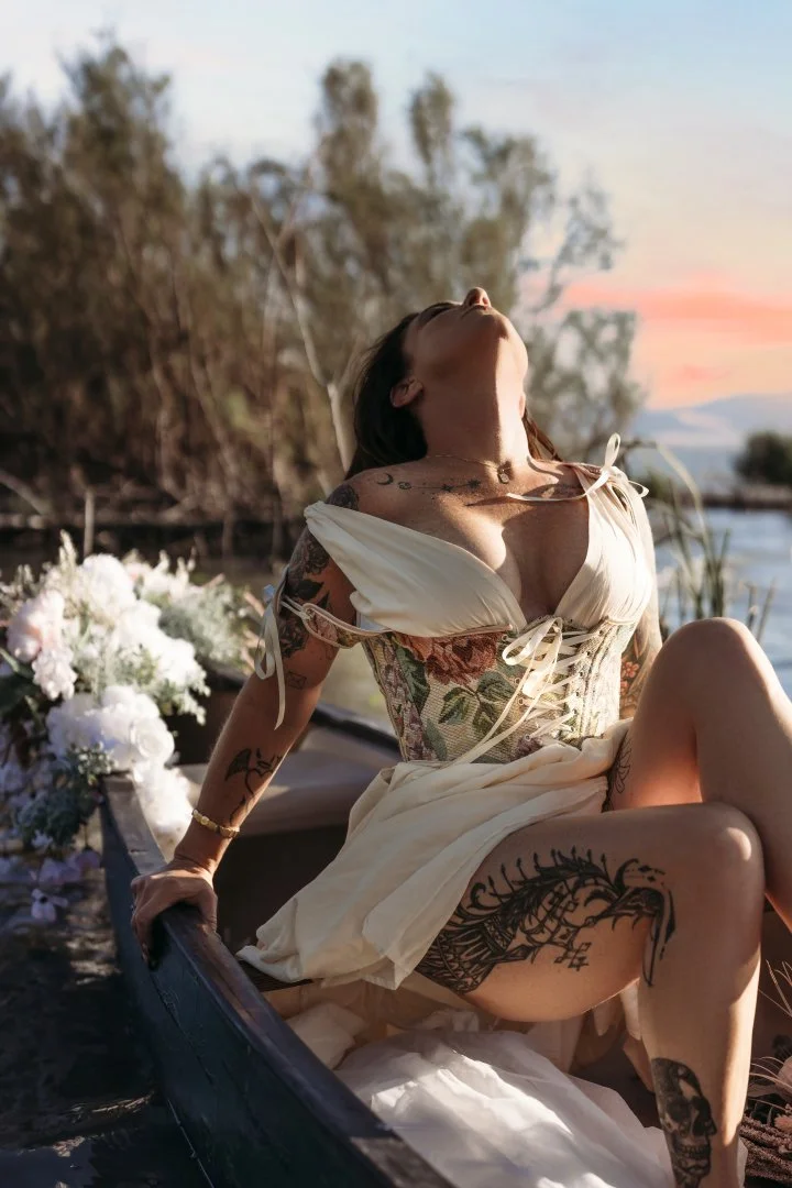 A woman with tattoos sitting in a boat near water at sunset, wearing a floral corset and a flowing cream-colored dress.