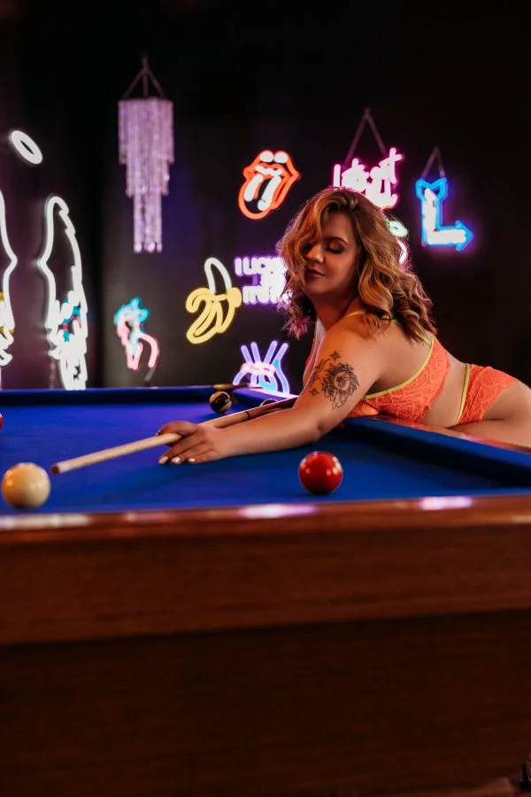 A woman with wavy hair and a tattoo on her arm is leaning over a pool table, aiming to hit a white cue ball with a pool cue in a dimly lit room with neon signs on the wall.