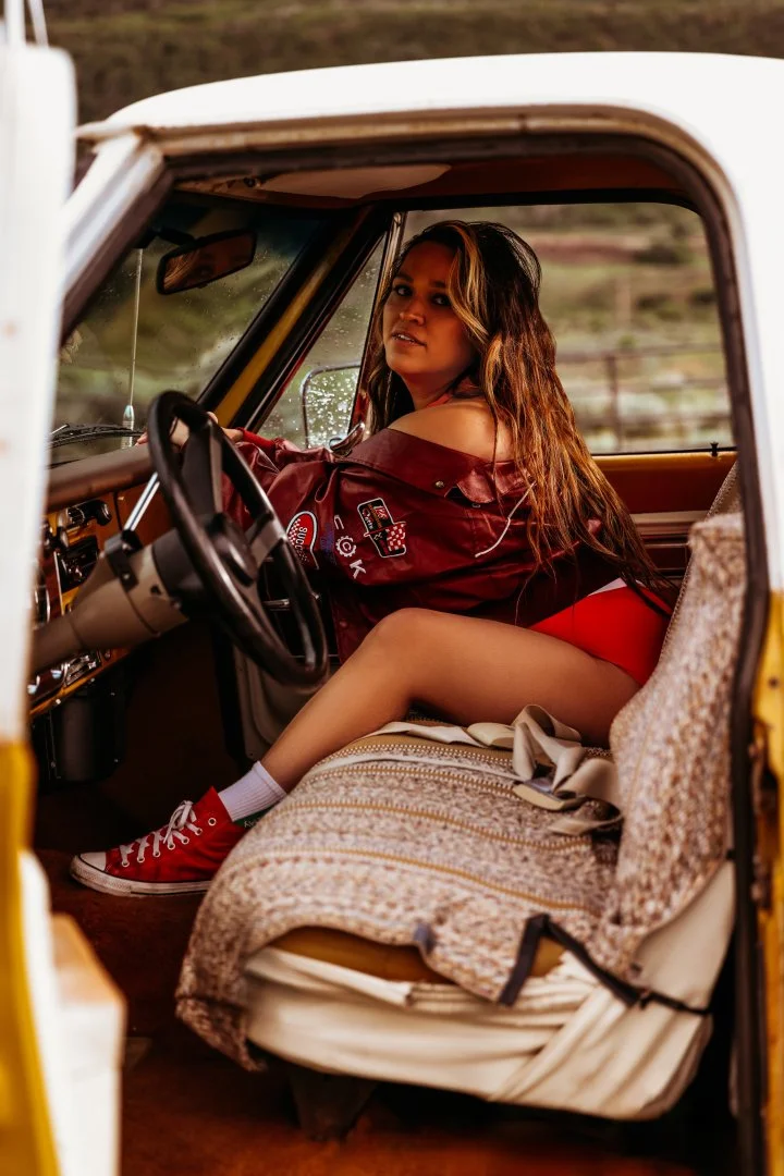 A young woman with long hair sitting in the driver's seat of a vintage truck, turning to look at the camera, with a rural landscape in the background.