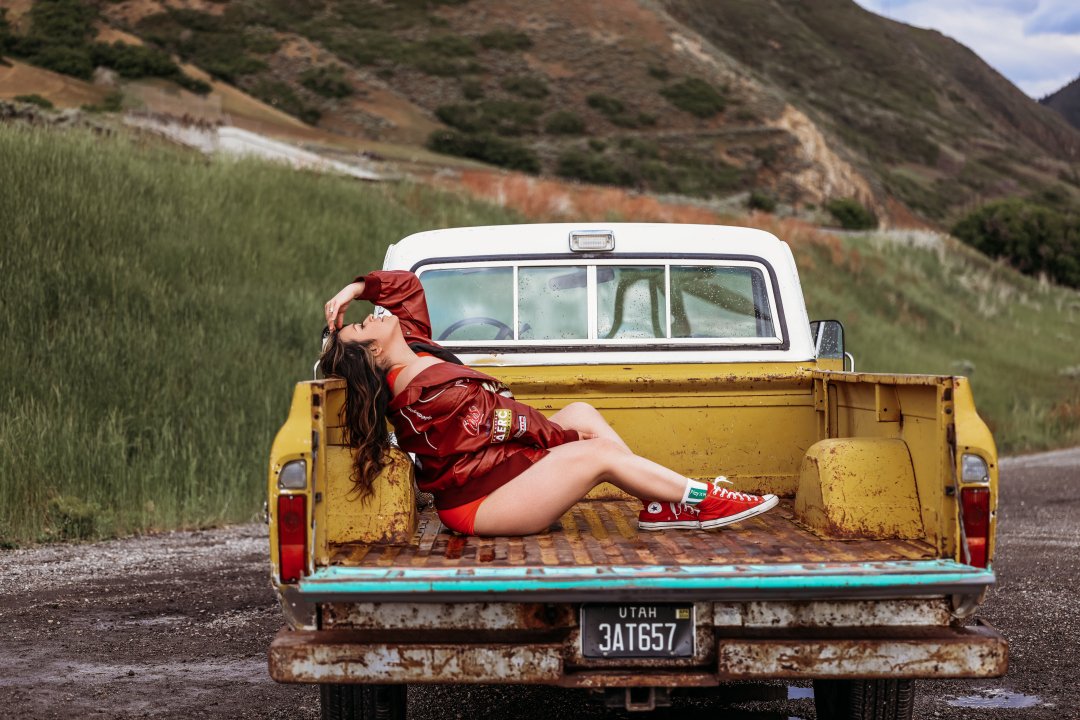Woman in red jacket and red sneakers lying in the bed of an old yellow pickup truck parked on a rural road, with green hills and mountains in the background.