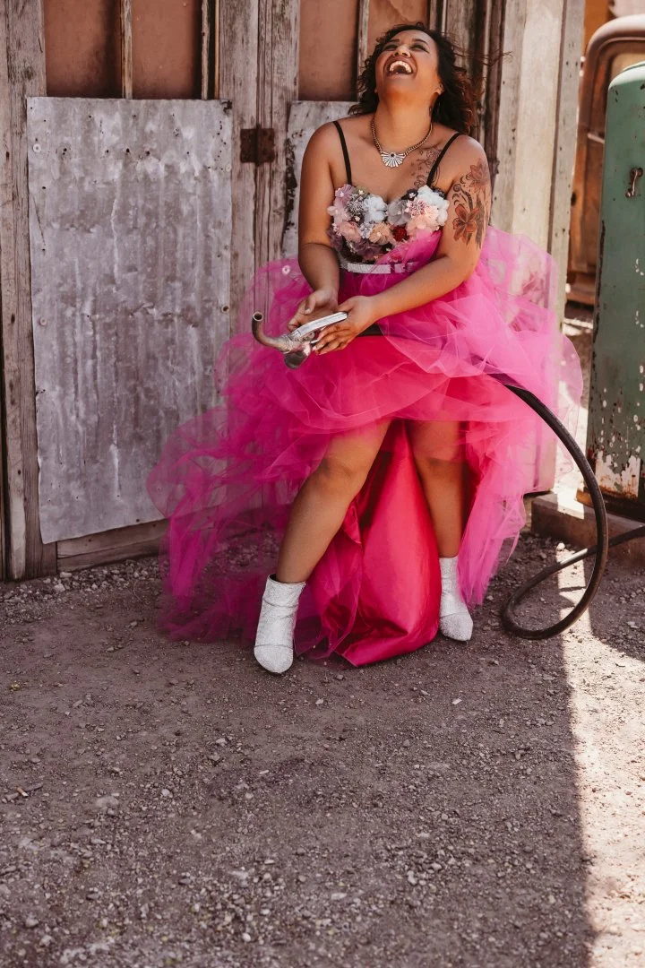 A woman with curly hair, tattooed arm, wearing a bright pink tulle dress, white high-heeled boots, and a beaded necklace, sitting outdoors on a pink stool, laughing and holding a silver horn, against a weathered wooden wall and rustic surroundings.