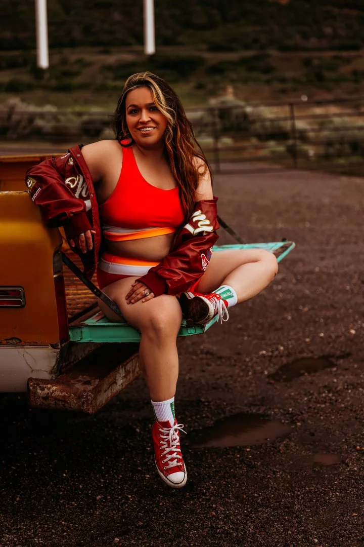 A woman sitting on the tailgate of a pickup truck in an outdoor setting, wearing a red sports bra, a red jacket, shorts, and red sneakers with white socks, smiling at the camera.