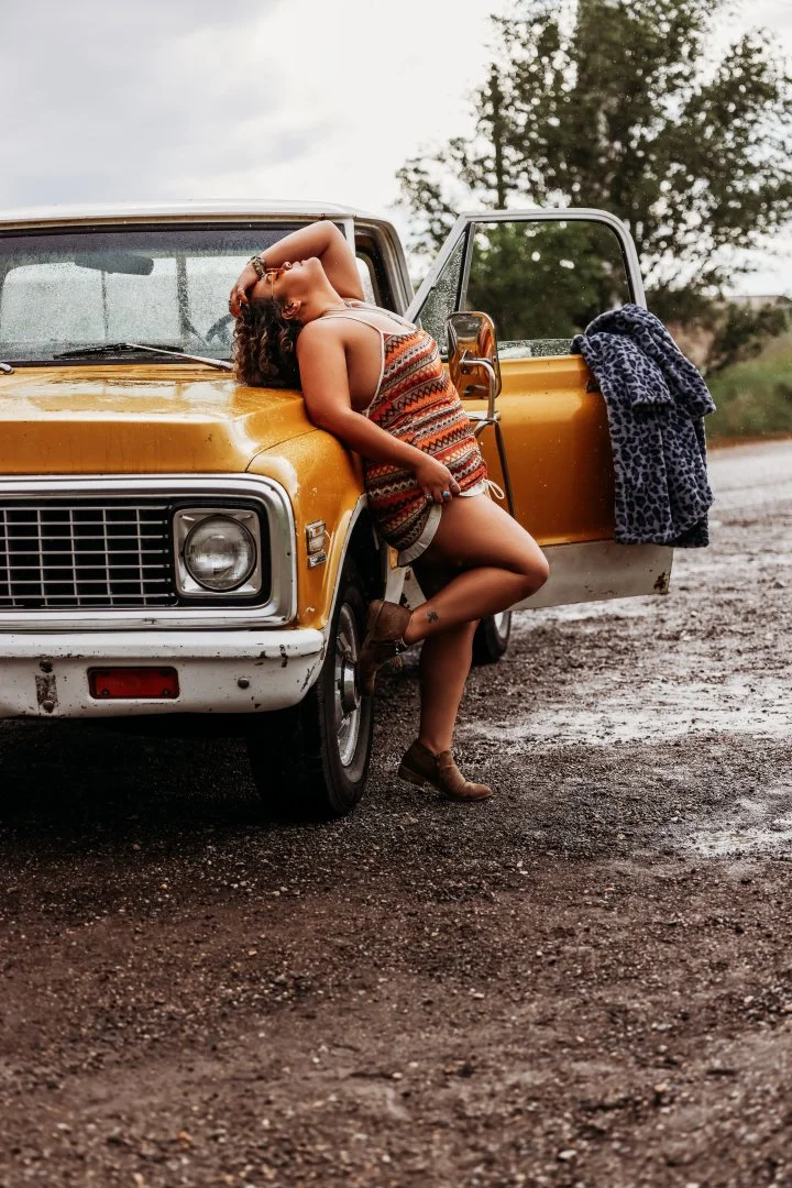 A woman with curly hair leaning against a vintage yellow truck on a dirt road, wearing a colorful patterned dress and brown boots, with a leopard print jacket hanging on the open door. Rain is falling, and trees are visible in the background.