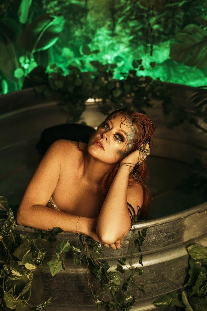 A woman with red hair and elaborate makeup relaxing in a vintage metal tub surrounded by green foliage with a green-lit forest background.