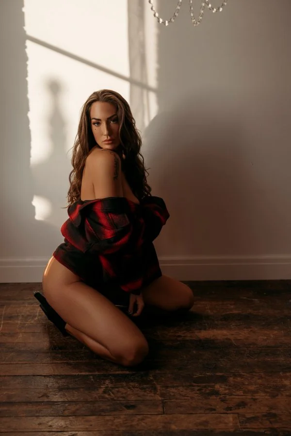 A woman kneeling on wooden floor with sunlight casting shadows on the wall, wearing an off-shoulder red and black plaid shirt.