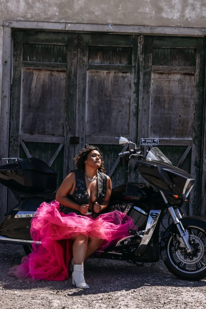 A woman is sitting on a black motorcycle in front of a weathered wooden barn door. She is wearing a vibrant pink tulle skirt, a black vest, and white boots. She has curly hair, a necklace, and appears to be posing with a confident expression.