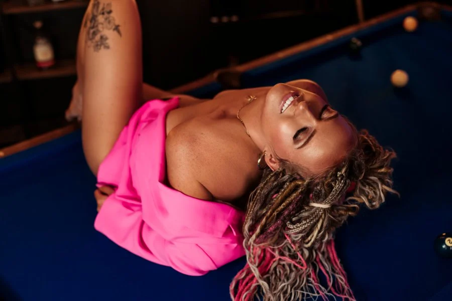 Woman with dreadlocks lying on a pool table, wearing a pink off-shoulder top, smiling with eyes closed.