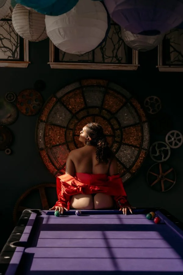 Woman in a red jacket sitting on a pool table with her back to the camera, holding pool cues, illuminated by soft lighting, with a large colorful circular wall art and movie reel decorations in the background.