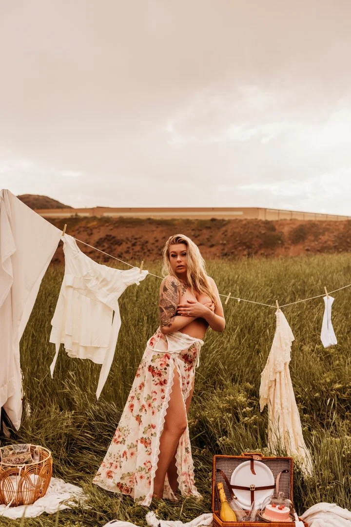 A woman with tattoos on her arm standing in a grassy field, holding a cloth to her chest, with laundry hanging on a line nearby and a picnic basket and blanket on the ground.