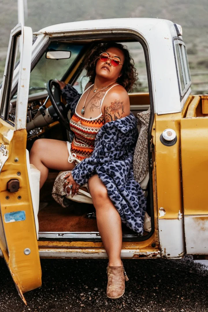 A woman with curly hair and sunglasses sitting in the driver's seat of a vintage yellow and white pickup truck, wearing a colorful patterned sundress, a blue animal print coat, and ankle boots, with a tattoo on her left shoulder and a necklace.
