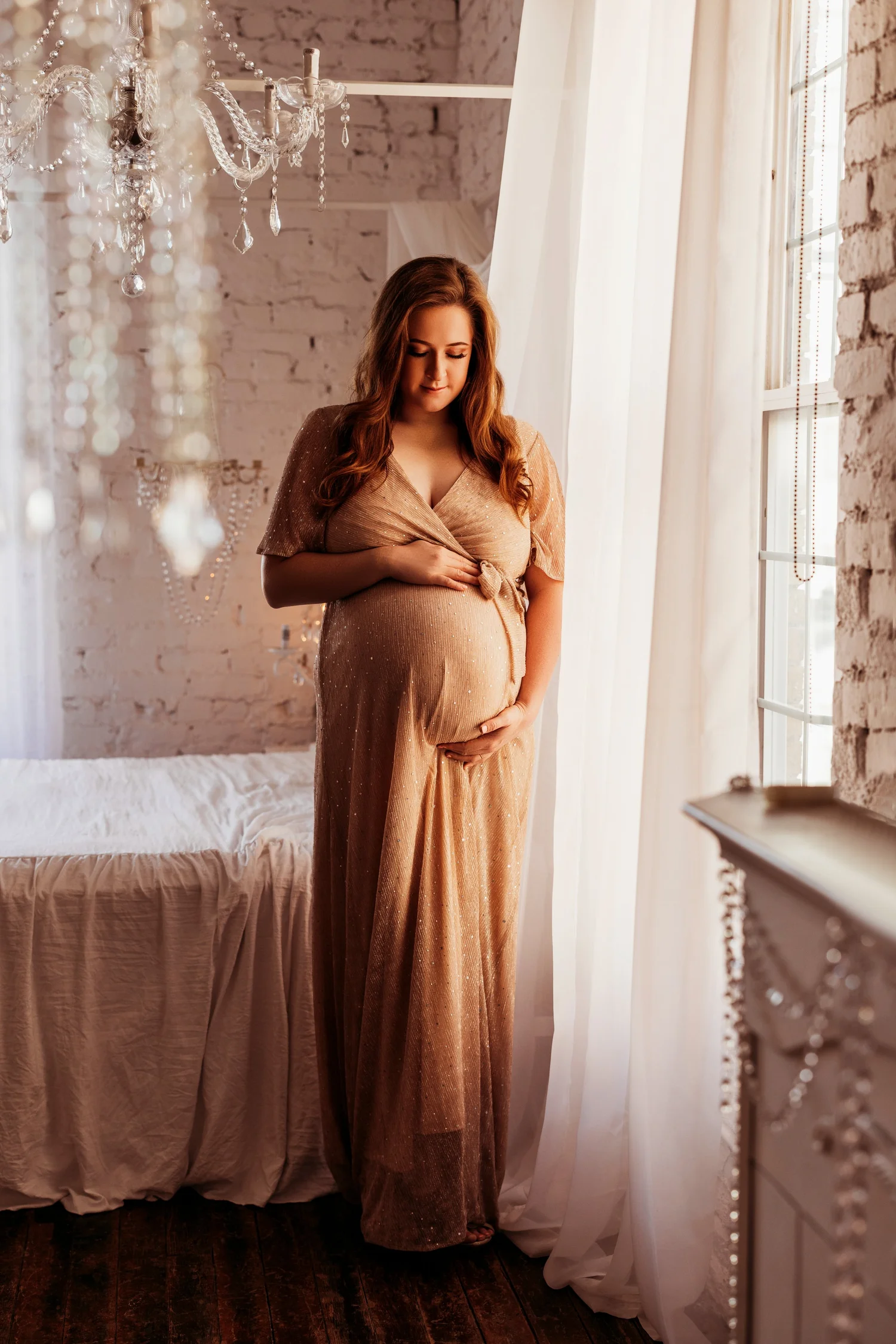A pregnant woman in a shimmering gold gown by a window with white curtains, cradling her belly