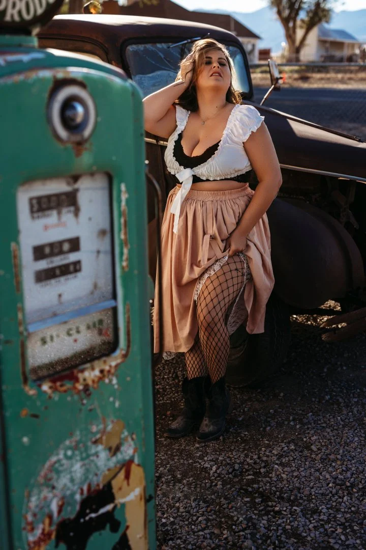 A woman with curly hair, wearing a white blouse with ruffled sleeves, pink skirt, fishnet stockings, and black boots, leaning against a vintage black truck next to an old, rusted green gas pump outdoors.