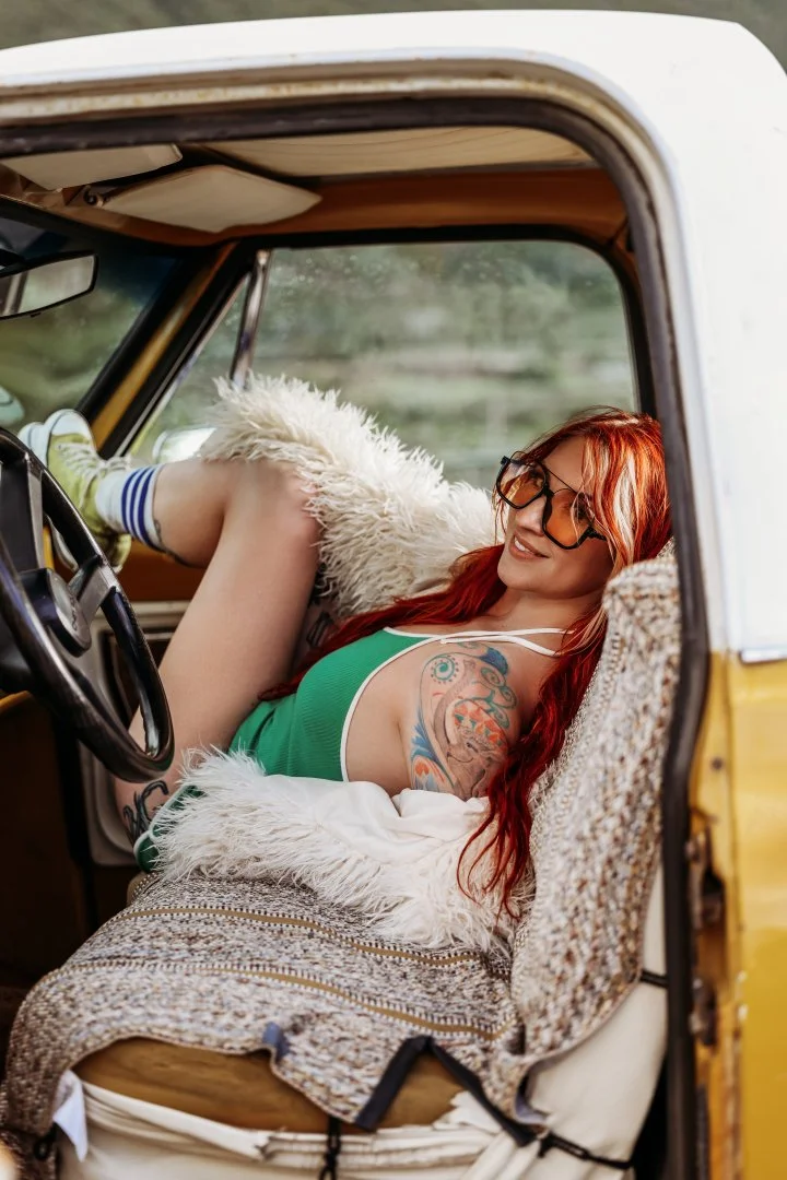 A woman with long red hair, glasses, and tattoos, wearing a green tank top and striped socks, relaxing inside an old vehicle with vintage decor.