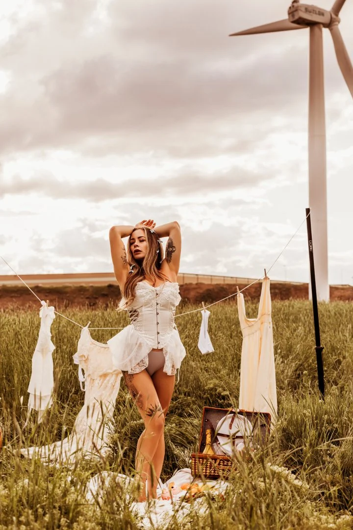 A woman with tattoos in a white top and gray underwear standing in a grassy field during daytime, with a wind turbine in the background and laundry hanging on a line in front of her. A picnic basket with bottles and plates is on the ground nearby.