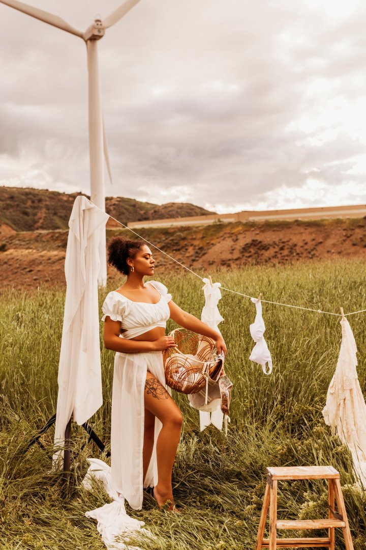 A woman standing outdoors in a grassy field, surrounded by a clothesline with white fabric items hanging to dry. A wind turbine is in the background, and the sky is overcast.