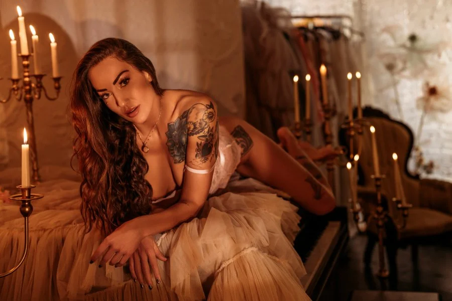 A woman with long wavy hair, tattoos, and makeup lying on a bed in a dimly lit room with candles.