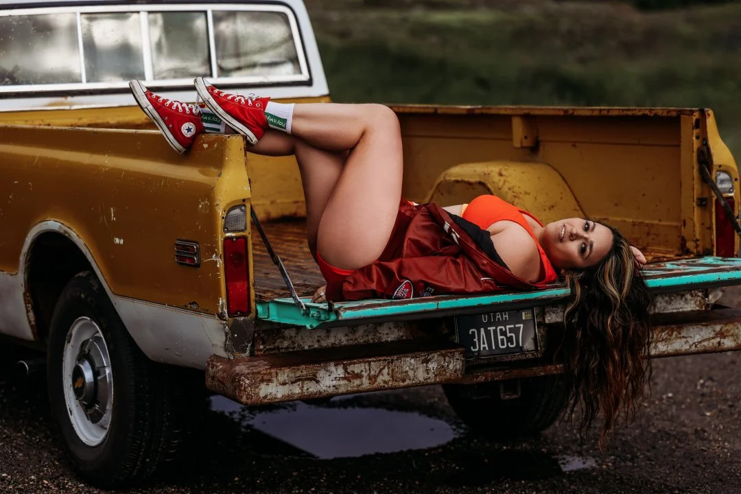 A woman with long, wavy hair lying on her side in the bed of a rusty, old yellow pickup truck with a Utah license plate. She is wearing red and black sneakers, a red satin jacket, and an orange top, posing outdoors with a blurred natural background.