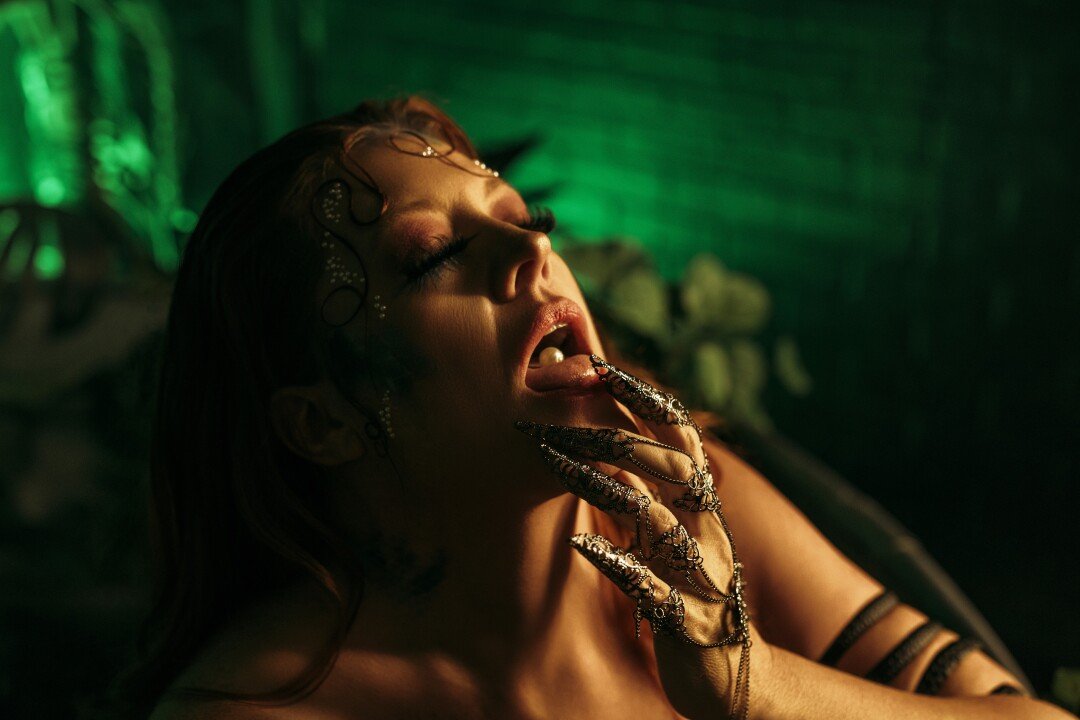A woman with dark makeup, multiple rings and jewelry covers her mouth with her gloved hand, tilting her head back with eyes closed in a dim green-lit setting.