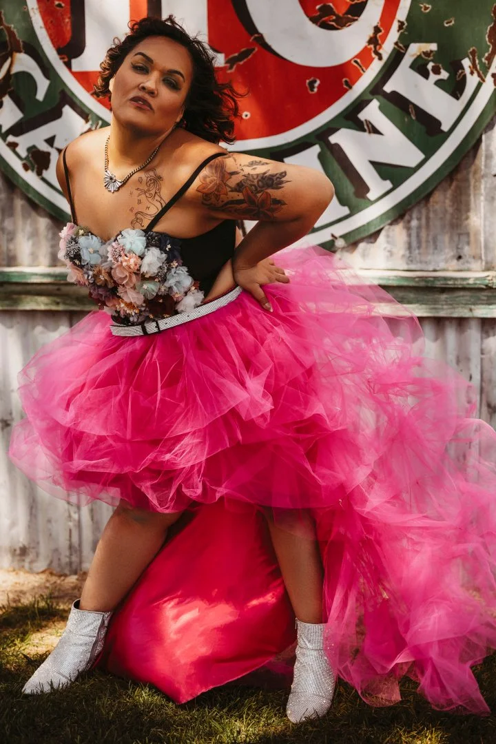 A woman with dark hair and tattoos wearing a pink tulle dress, silver boots, and a floral top, standing in front of a weathered fence with a backlit logo.