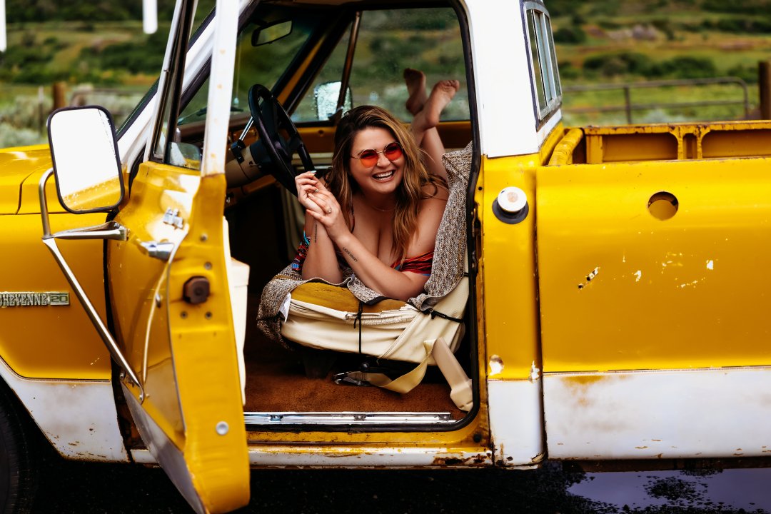 A woman with long hair, sitting inside a yellow and white vintage truck, smiling and wearing sunglasses with a cheerful expression.