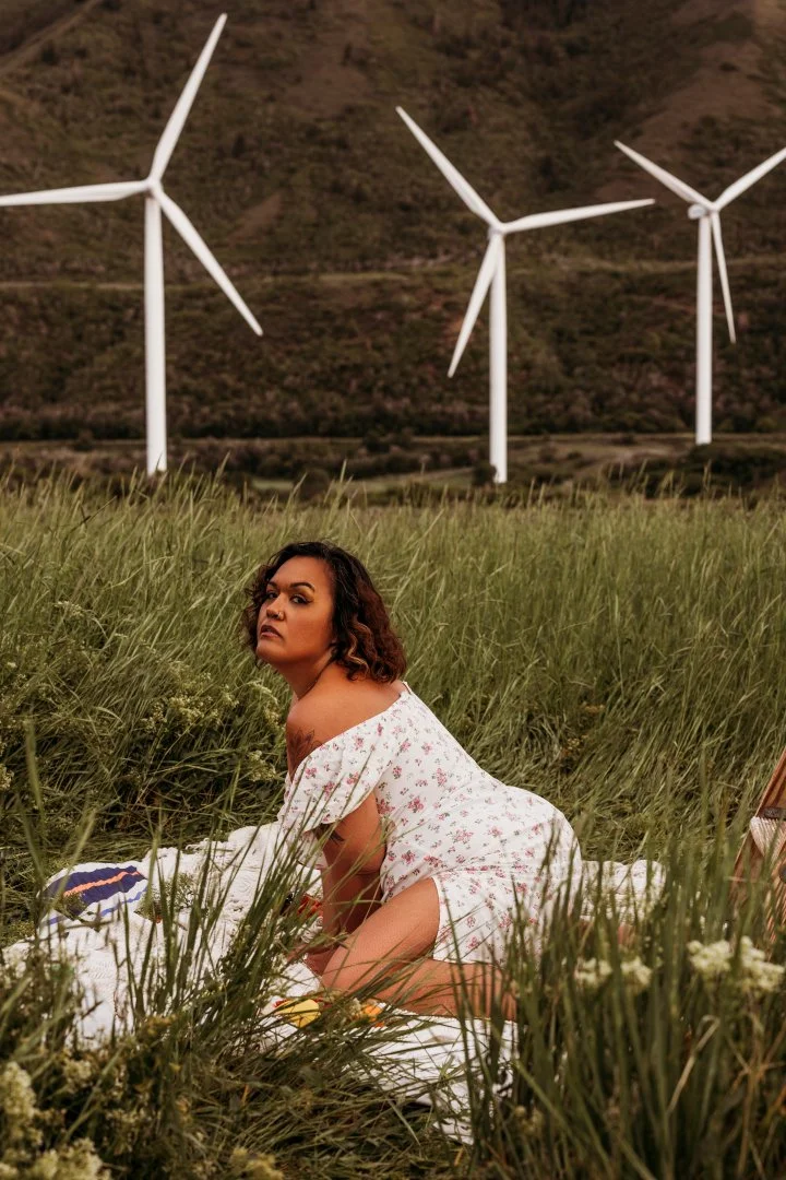 A woman in a floral dress kneeling in tall grass with wind turbines in the background.