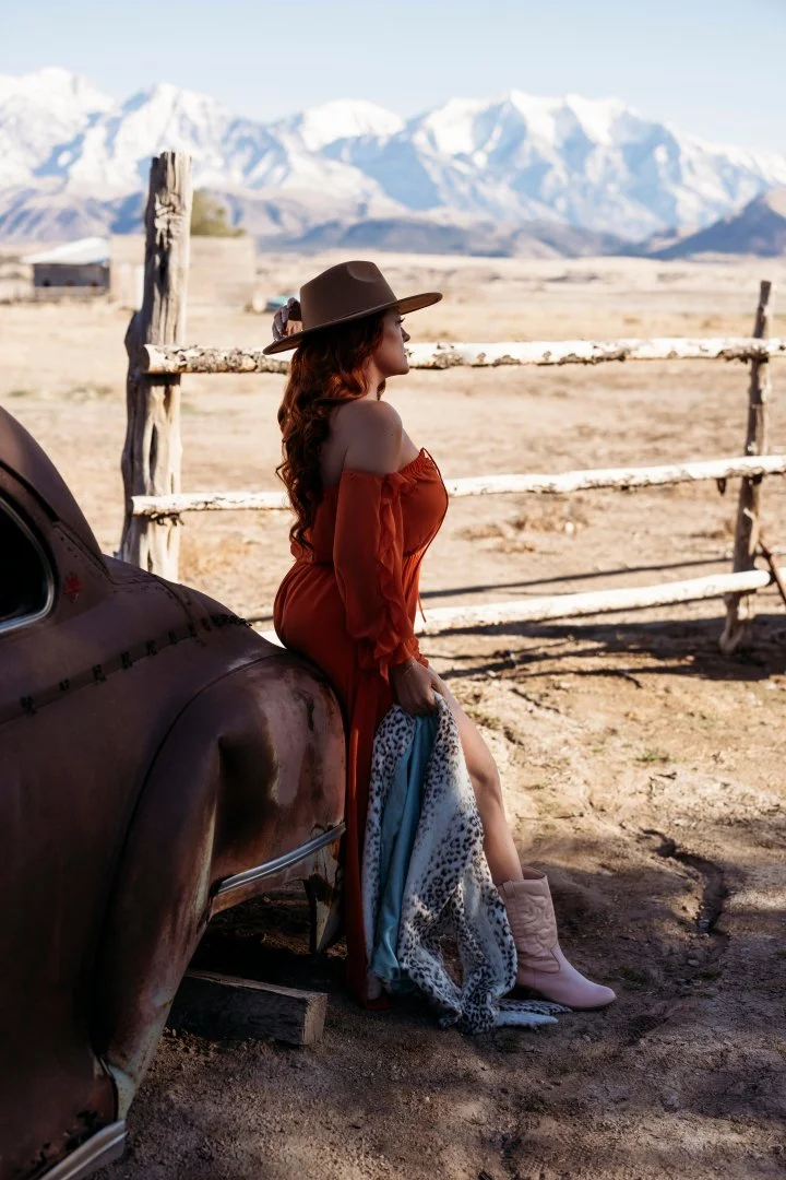 A woman in a burnt orange off-shoulder dress and cowboy hat sitting on the fender of an old rusty car, holding a patterned scarf, with a rustic fence, desert landscape, and snow-capped mountains in the background.