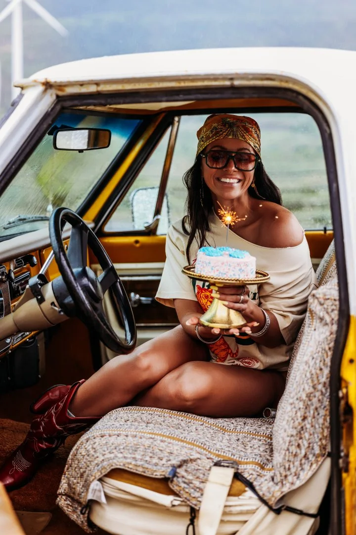 Woman smiling inside a vintage yellow truck, holding a birthday cake with a sparkler on top, wearing sunglasses and a bandana.