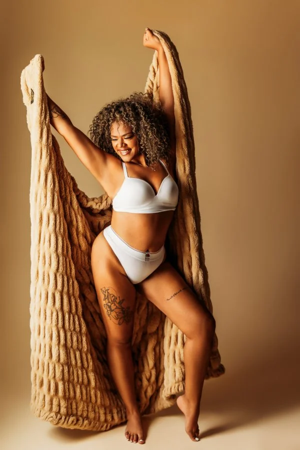 A woman with curly hair wearing white lingerie, standing against a beige background, holding a large beige blanket or faux fur coat, smiling with arms raised.
