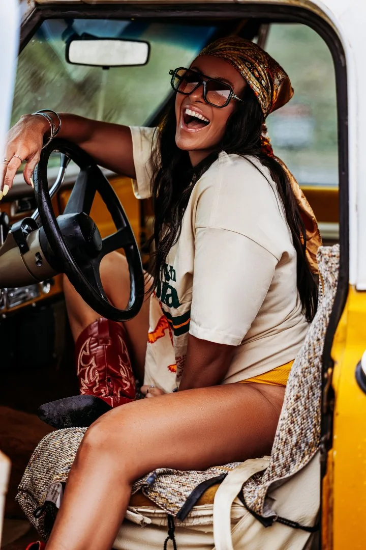 Woman wearing glasses and a headscarf sitting in the driver's seat of a yellow vehicle, smiling and laughing.