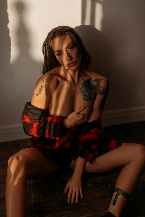 Young woman with tattoos sitting on the floor in a dimly lit room, wearing a red and black flannel shirt partially off her shoulders, with shadows cast on the wall behind her.