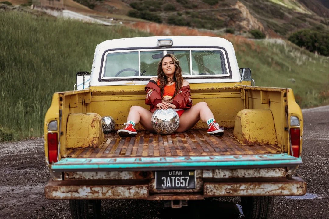 A woman sitting in the back of a yellow pickup truck with a wooden bed, holding a disco ball, with two other disco balls beside her. She is outdoors on a rural road with green hills and mountains in the background.