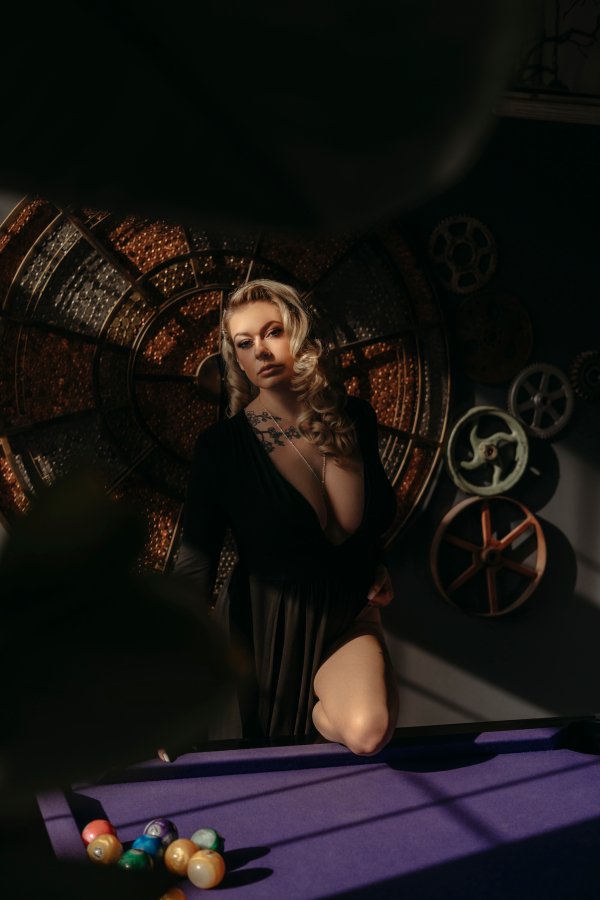 A woman with blonde curly hair and makeup posing near a pool table, with a dark background featuring film reels and circular lights.