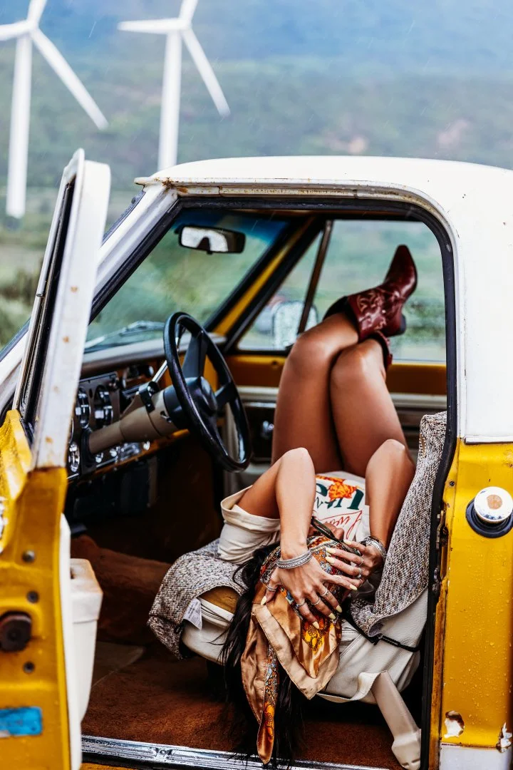 Person with long dark hair, colorful jewelry, and patterned scarf resting inside a vintage yellow and white car with open door, holding their legs and sitting with knees up and head down.