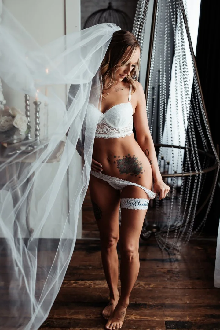 A bride in white lace lifting a garter that reads Bride, amid floral decor