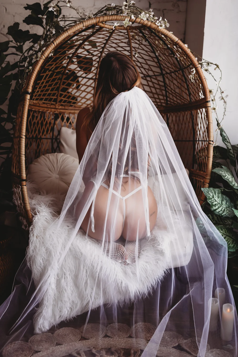 A bride in white lingerie and a veil kneeling within a wicker canopy with florals