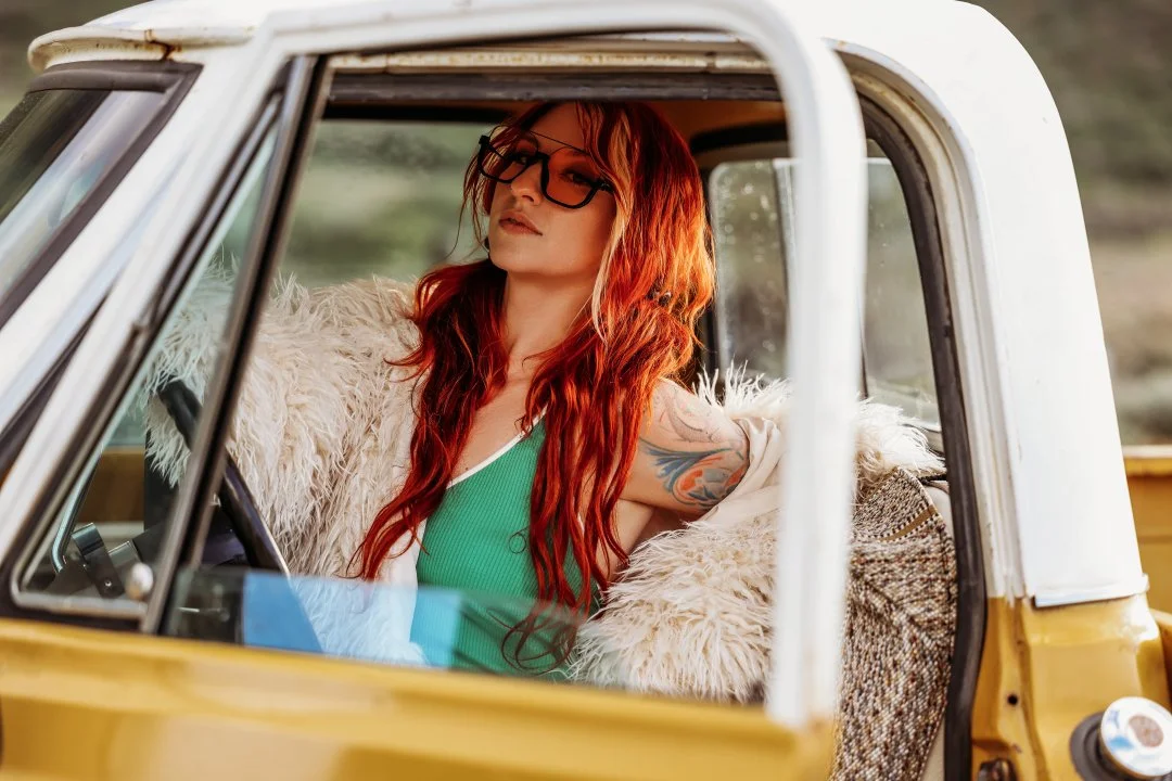 A woman with long red hair, glasses, and tattoos inside an old yellow and white vehicle, looking out the window with a relaxed expression.