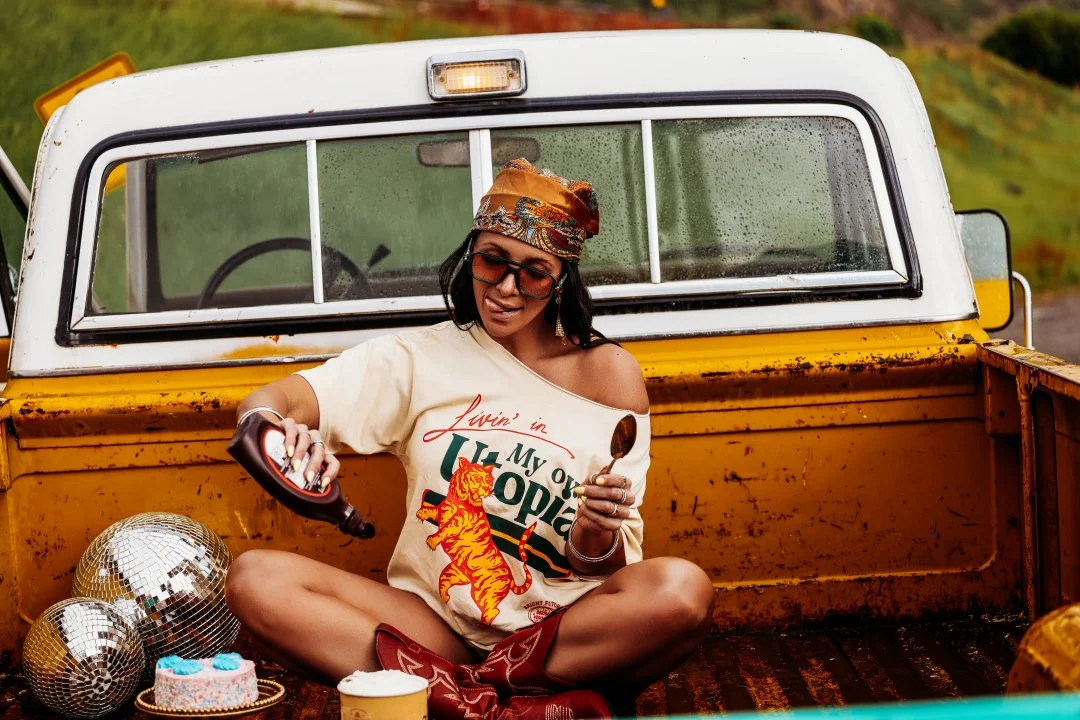 A woman sitting cross-legged in the bed of a rusty yellow pickup truck, pouring a drink, with disco balls and a birthday cake nearby. She is wearing sunglasses, a patterned headscarf, a T-shirt with a tiger and the text 'Livin’ in Uganda, My old Otop