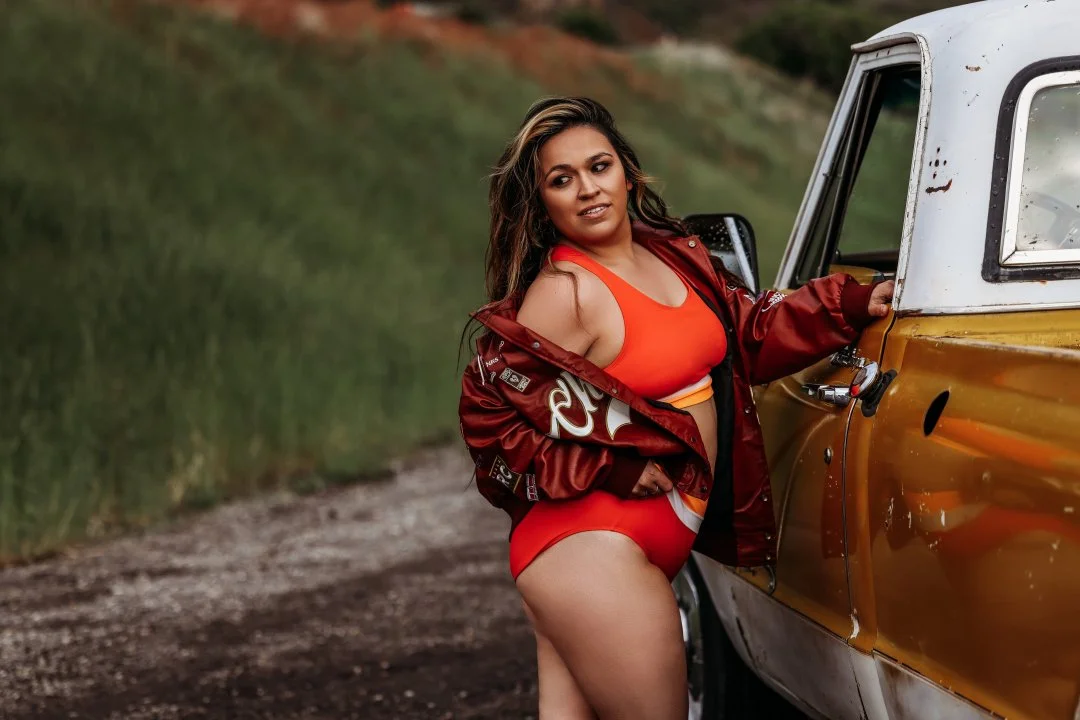 Woman in red sports bra and shorts standing next to a vintage yellow truck outdoors.
