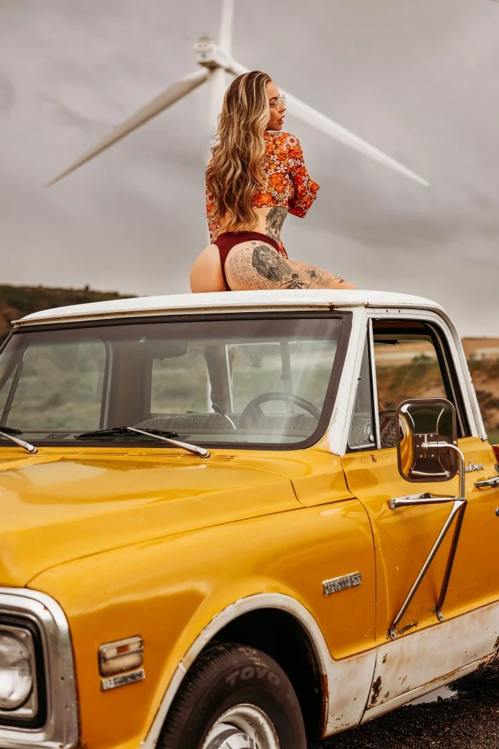 A woman with long wavy hair and colorful tattoos sitting on the roof of a yellow vintage truck, with a wind turbine in the background and a cloudy sky.