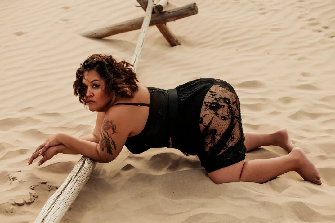 Woman with curly hair and tattoo on arm wearing black lace dress on sandy beach, leaning on a wooden pole.