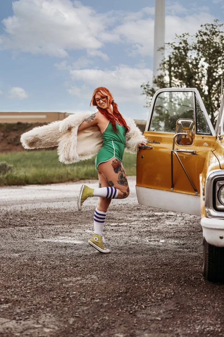 Woman with red hair and tattoos, wearing a green dress, white jacket, knee-high socks, and yellow sneakers, smiling and holding open a yellow vintage van door outdoors against a partly cloudy sky.
