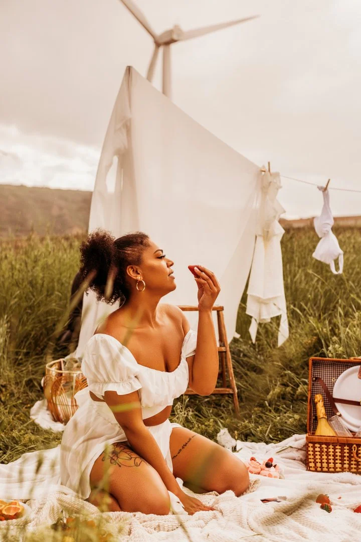 A woman with dark curly hair sitting on a picnic blanket in a field with tall grass. She is wearing a white off-shoulder dress with puffed sleeves. Behind her, there's a large white sheet hanging from a clothesline and a wind turbine in the distance.