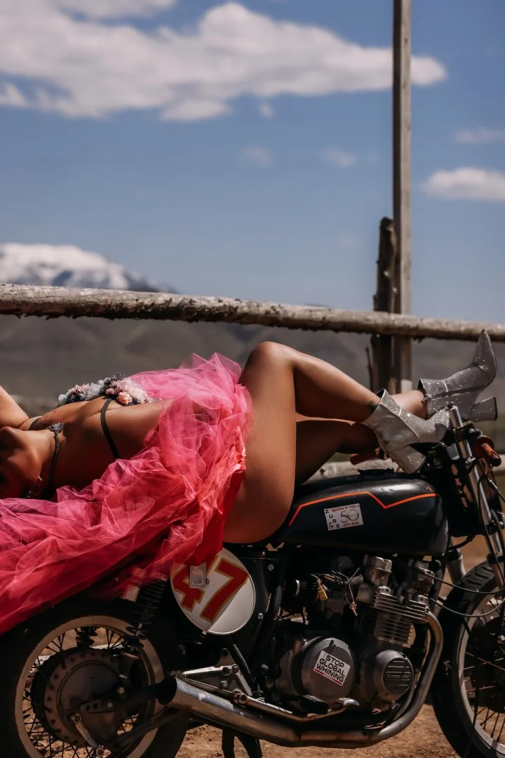 A woman lying on a black motorcycle with pink tulle skirt, silver boots, and a flower headband in an open landscape under a blue sky with clouds.
