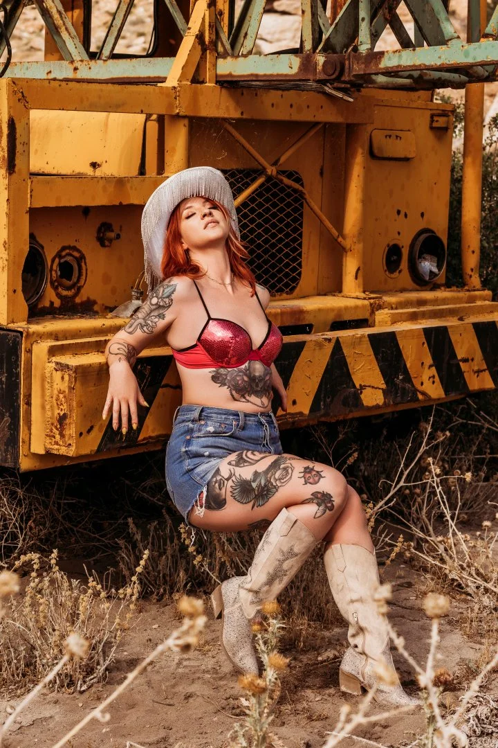 A woman in a wide-brimmed hat and sequined top leaning on a rusted construction vehicle