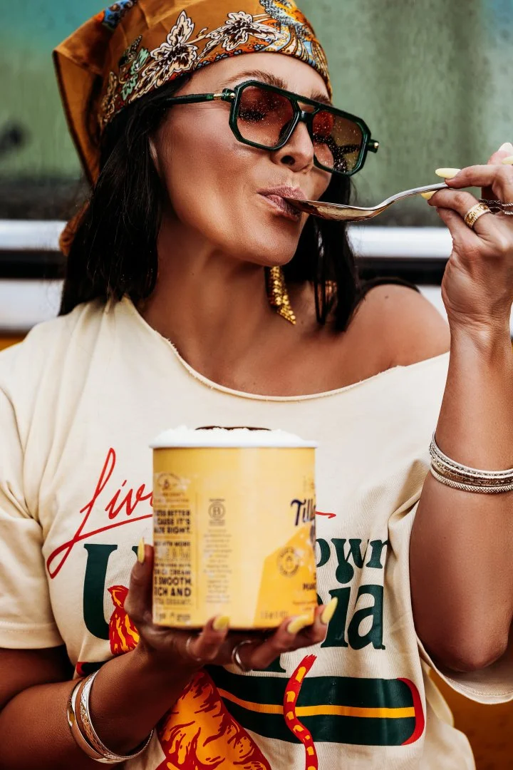 Woman wearing large sunglasses and a colorful headscarf, holding a spoon with ice cream close to her lips and a container of ice cream in her other hand.