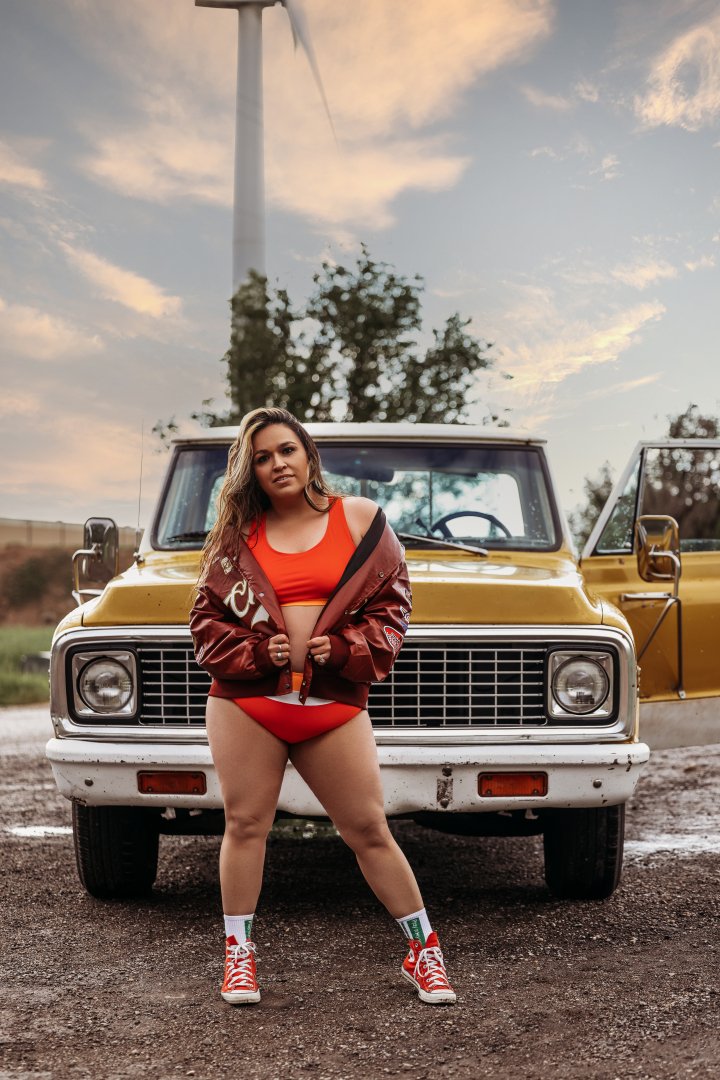 A woman in red sports bra, red shorts, and a maroon jacket standing in front of a yellow vintage SUV with a wind turbine in the background during sunset.