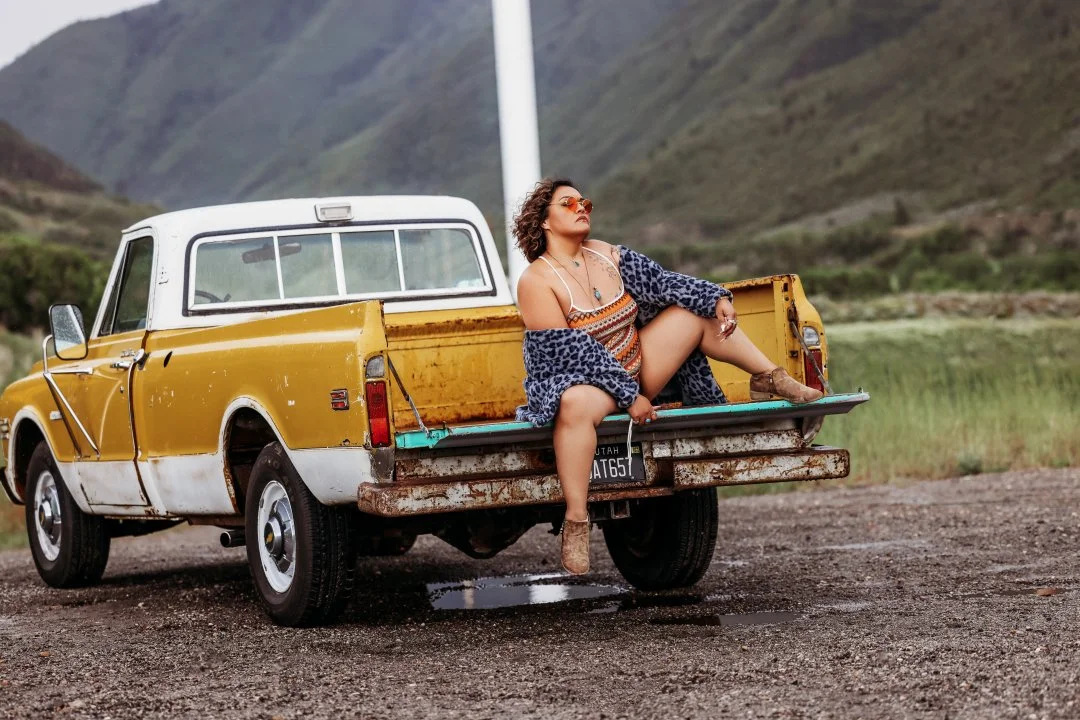 A woman sitting on the tailgate of a vintage yellow pickup truck in a rural landscape with mountains in the background.